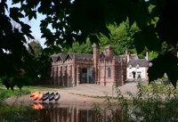 A brick building with crenellations and towers with a house and blue sky in the background and canoes on a riverbank in the foreground.