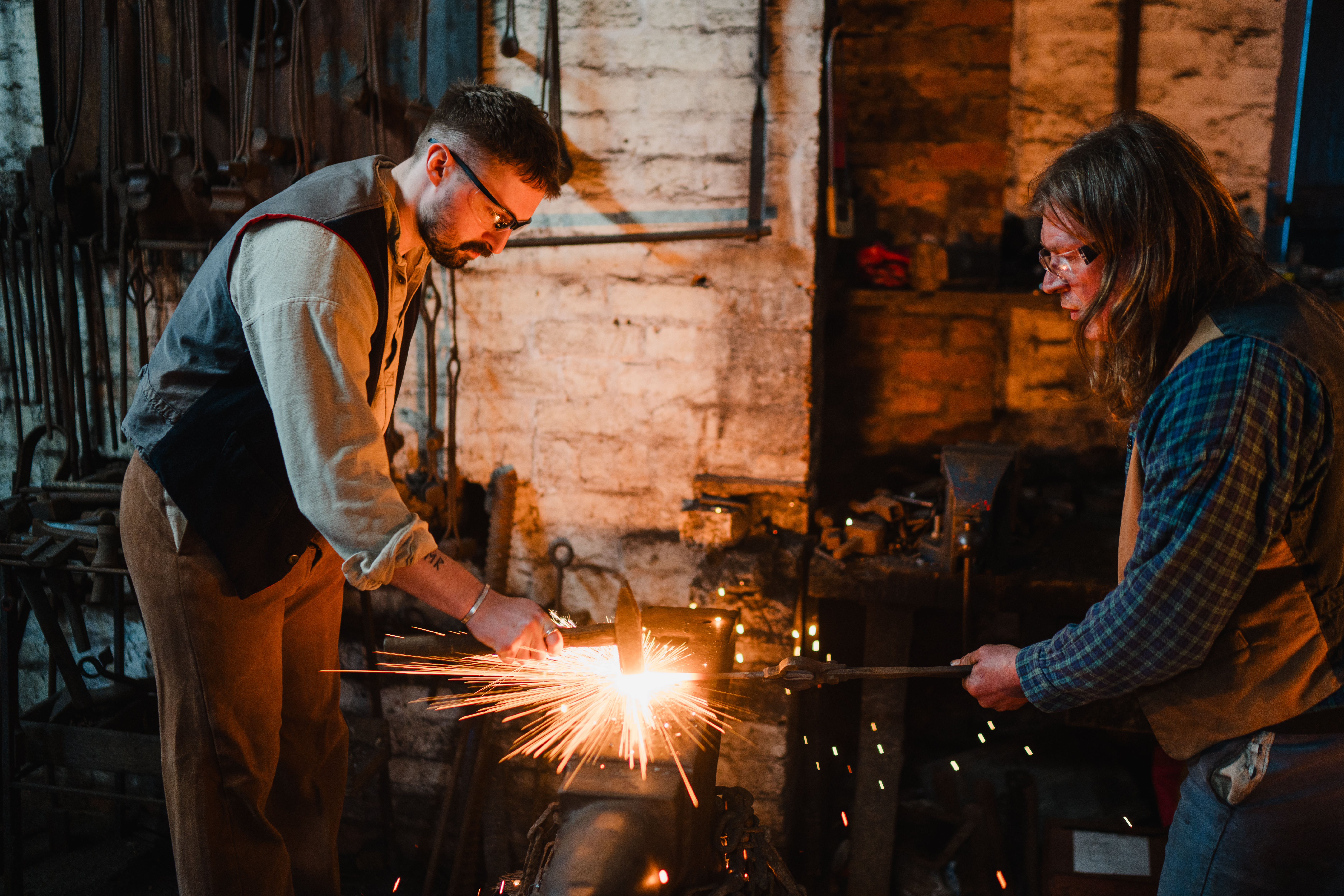 A young man, dressed in Victorian costume, is hitting a red-hot iron bar with a hammer on an anvil. There are sparks flying off the bar. They are in a blacksmith's forge.