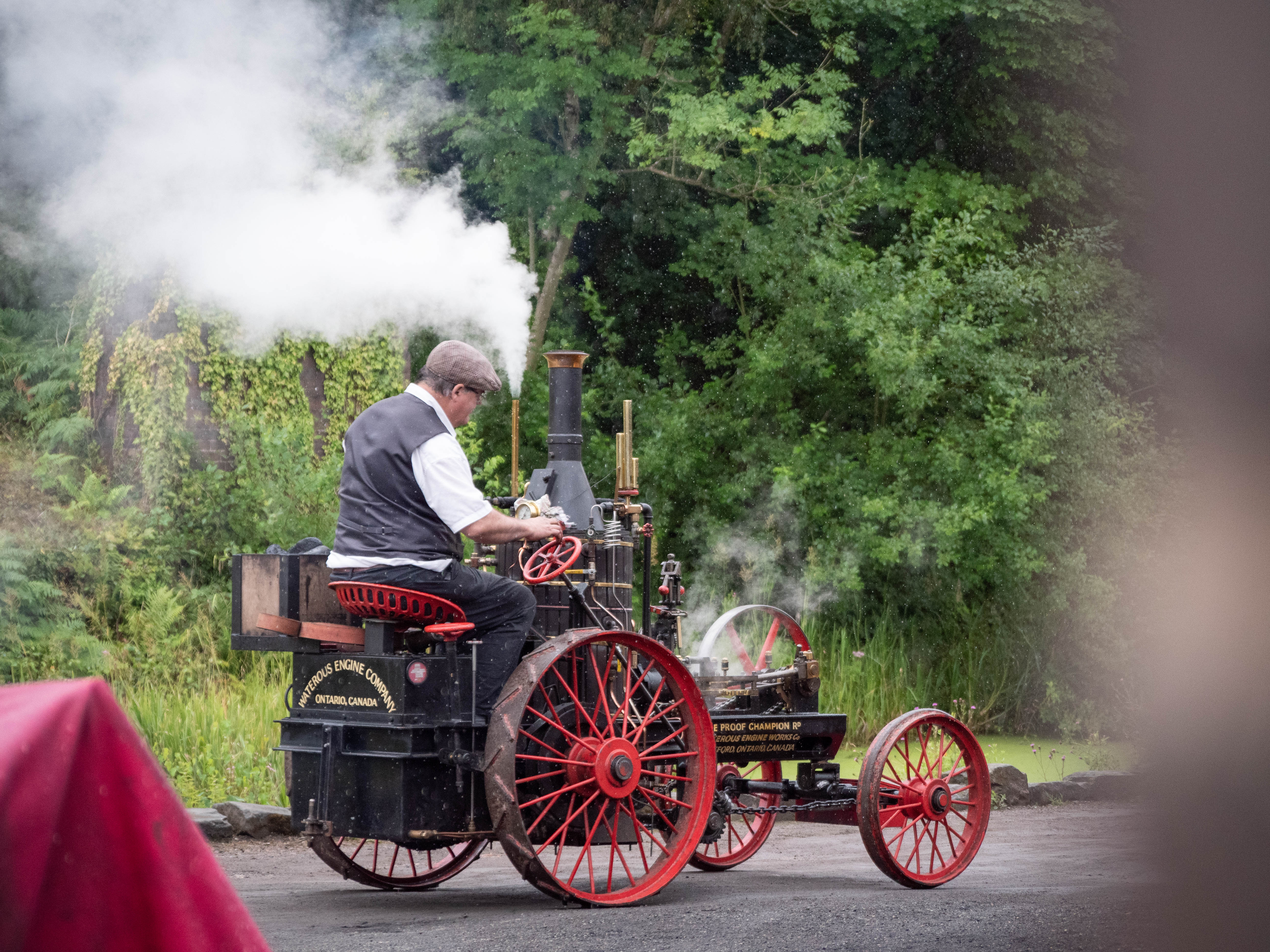 A man in Victorian costume is driving a black and red steam engine alongside a wooded canal towpath. There is smoke coming from the brass chimney on the engine.